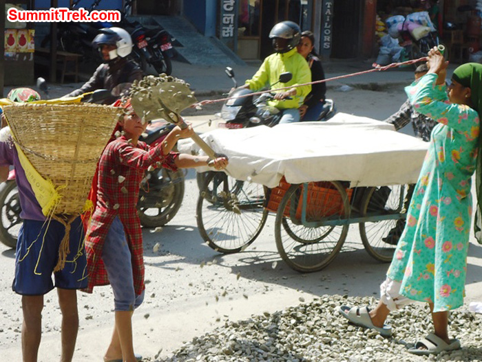 Kathmandu - motorcyclists drive past, while a peddlers rolls a fruit cart covered with a white sheet. 2 women load basket with gravel, one with shovel and other aiding with rope tied to shovel. Photo Carla Strong